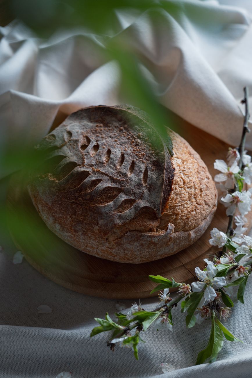 Rustic sourdough bread with intricate scoring, placed on a wooden board, surrounded by soft spring blossoms.