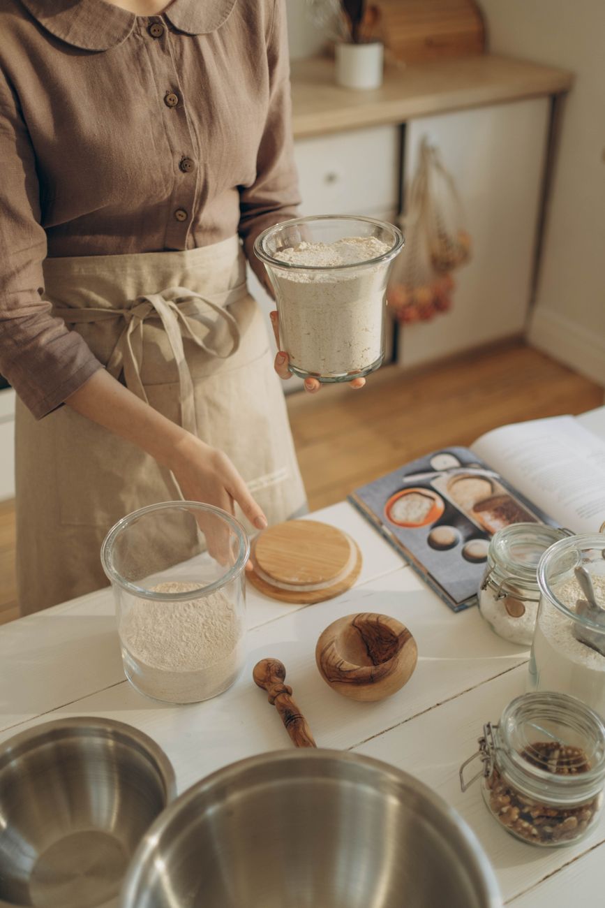 A woman measures flour for baking in a warm, inviting kitchen setting, surrounded by utensils.